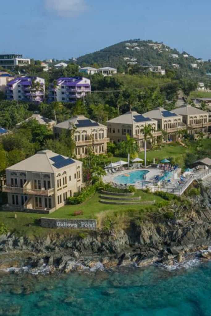 Aerial view of Gallows Point in St. John, USVI, with beige buildings, a pool, and lush greenery overlooking the ocean. Hills and additional structures entice visitors to explore St. John USVI activities and enjoy the stunning scenery.