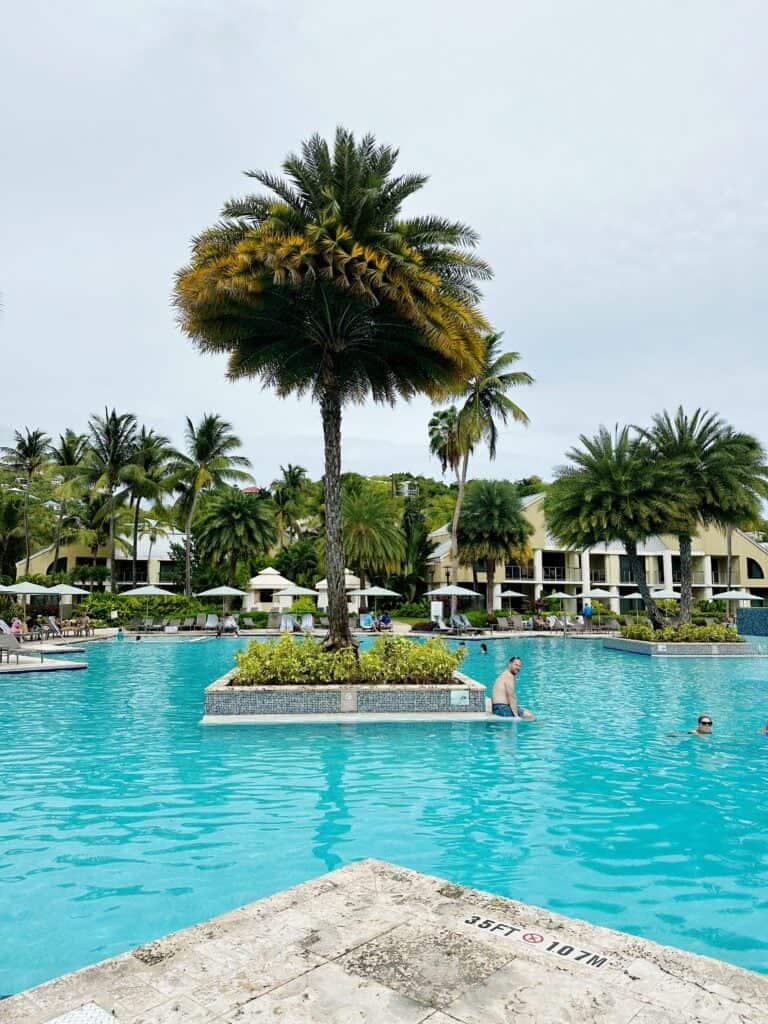 The outdoor swimming pool, a popular St. John USVI activity, boasts a central island featuring a palm tree. Visitors swim and relax around the pool, with buildings and more palm trees in the background under a cloudy sky—an iconic thing to do in St. John U.S. Virgin Islands.