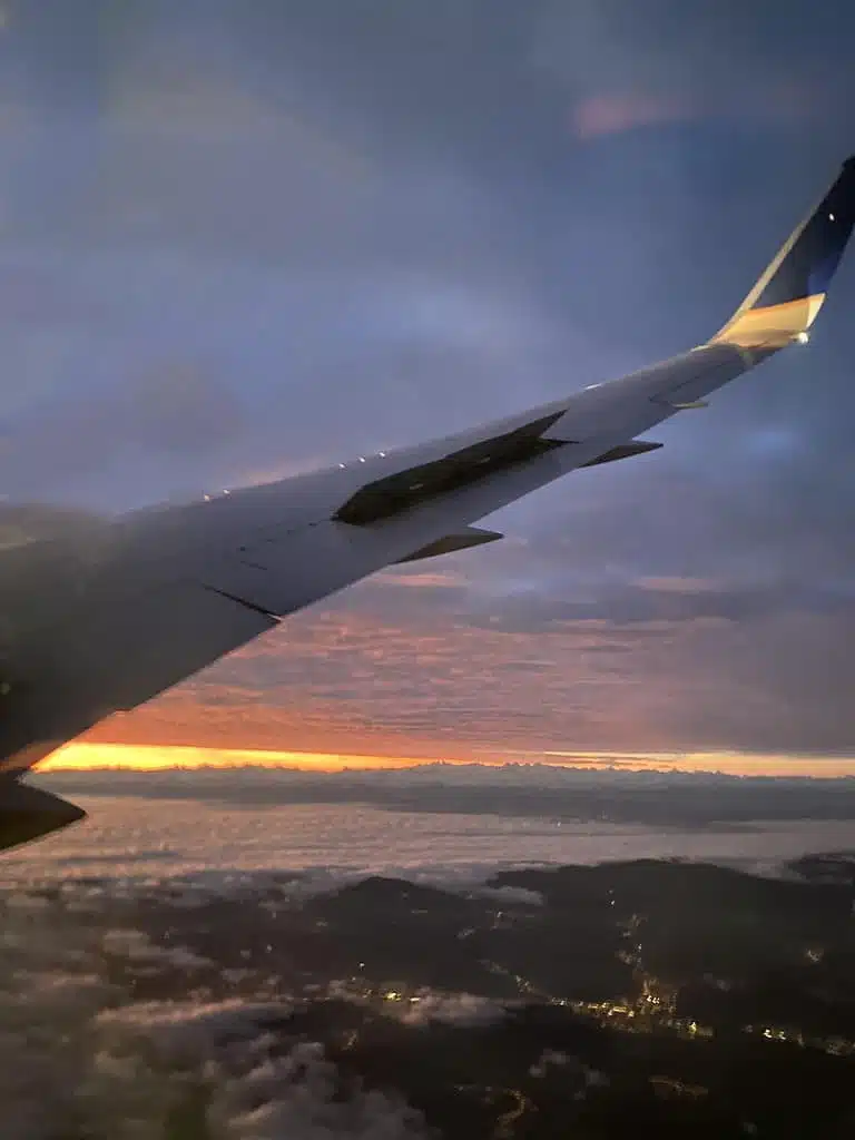 View from an airplane window showing the wing against a backdrop of a sunset sky, with clouds and lit-up cityscape below—an enchanting end to surviving a long-haul flight.