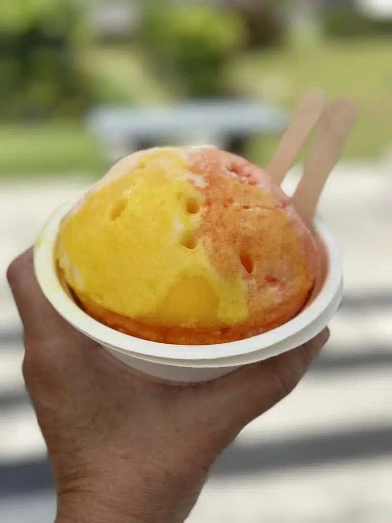 A hand holds a cup of shaved ice with orange and yellow flavors, perfect for a Maui itinerary 5 days trip. Two wooden spoons are tucked into the icy treat, ready to enjoy. The background is blurred.