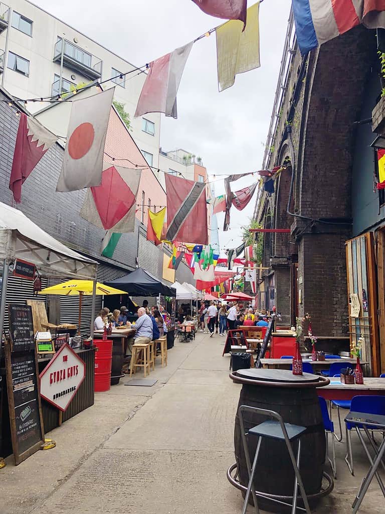 An outdoor market with various flags hanging above, people sitting at tables, and food stalls along a narrow alleyway beside a brick building.