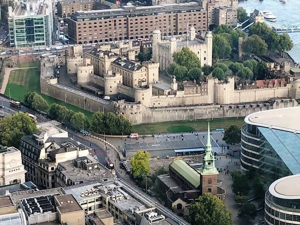 Aerial view of the Tower of London surrounded by trees and historical buildings with city architecture in the background. Several vehicles and people are visible near the site.