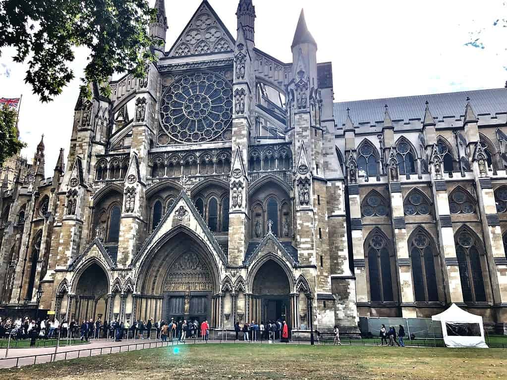 Visitors lining up outside the majestic Westminster Abbey in London, with its Gothic architecture featuring the large rose window and intricate stonework.