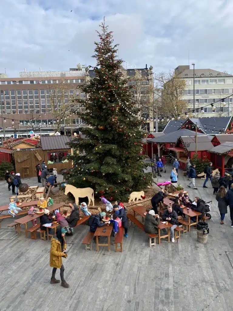 The Christmas tree at Wienachtsdorf Christmas Market at Sechelautenplatz. There are rustic chalets and tables around the tree to eat and drink Glühwein. 