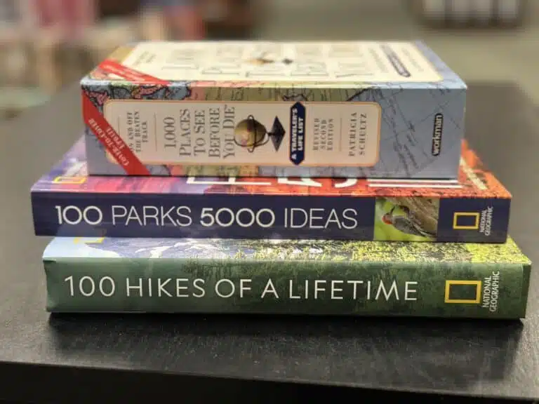 A stack of books for travel lovers sits on the table, featuring "1,000 Places to See Before You Die," "100 Parks 5000 Ideas," and "100 Hikes of a Lifetime," all published by National Geographic.