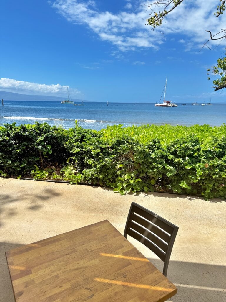 A wooden table and black chair on a patio overlook the ocean with sailboats in the distance and greenery along the shore—an inviting scene reminiscent of West Maui restaurants under a partly cloudy sky.