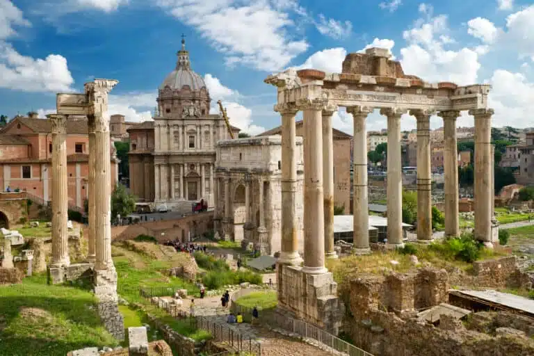 Ancient Roman ruins stand proudly under a blue sky with fluffy clouds, showcasing a rich tapestry of history. The foreground features towering Corinthian columns while the background reveals the grandeur of the Roman Forum, with visitors exploring the historic site.