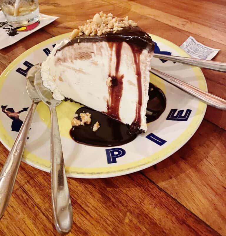 A delectable slice of chocolate drizzled pie with fluffy whipped cream topping and a crunchy nut garnish, served on a plate with 'HULA PIE' design, accompanied by a dessert spoon, ready for a sweet indulgence.