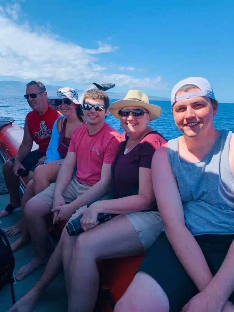 "Group of five people, with three women and two men, smiling and seated on a boat with clear blue skies and calm ocean waters in the background, featuring a whale mid-leap in the distance."