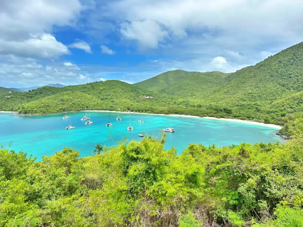 An aerial view of a scenic bay with turquoise water, several anchored boats, and lush green hills under a partly cloudy sky.