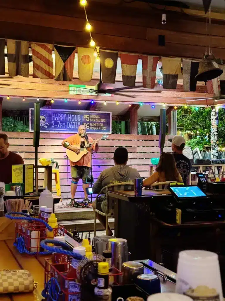 A musician plays acoustic guitar on an outdoor stage under string lights in a bar. Patrons are seated at the bar and tables, and a sign advertising happy hour is visible in the background.
