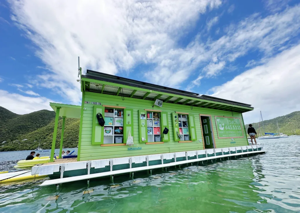 A green houseboat with an array of items displayed on its wall is anchored in clear water, against a backdrop of hills and a partly cloudy sky.