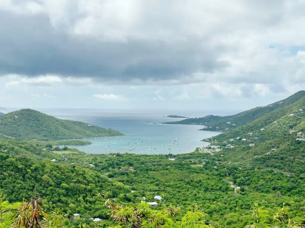 A lush, green landscape leading to a bay filled with numerous small boats on a cloudy day, surrounded by rolling hills and partially covered by a cloudy sky.