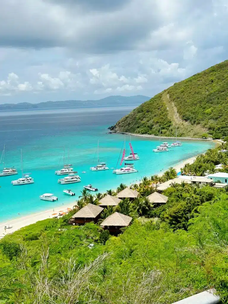 Elevated view of a coastal scene featuring a turquoise bay with sailboats, lush green hillside, sandy beach, and thatched-roof huts amidst dense foliage under a partly cloudy sky.