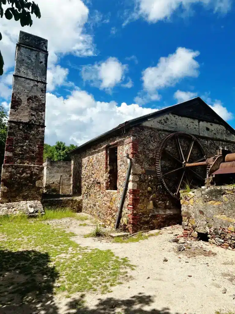 Old stone building with a large wheel mounted on one wall, a tall chimney, and remnants of brick structures around it, surrounded by greenery under a blue sky with scattered clouds.
