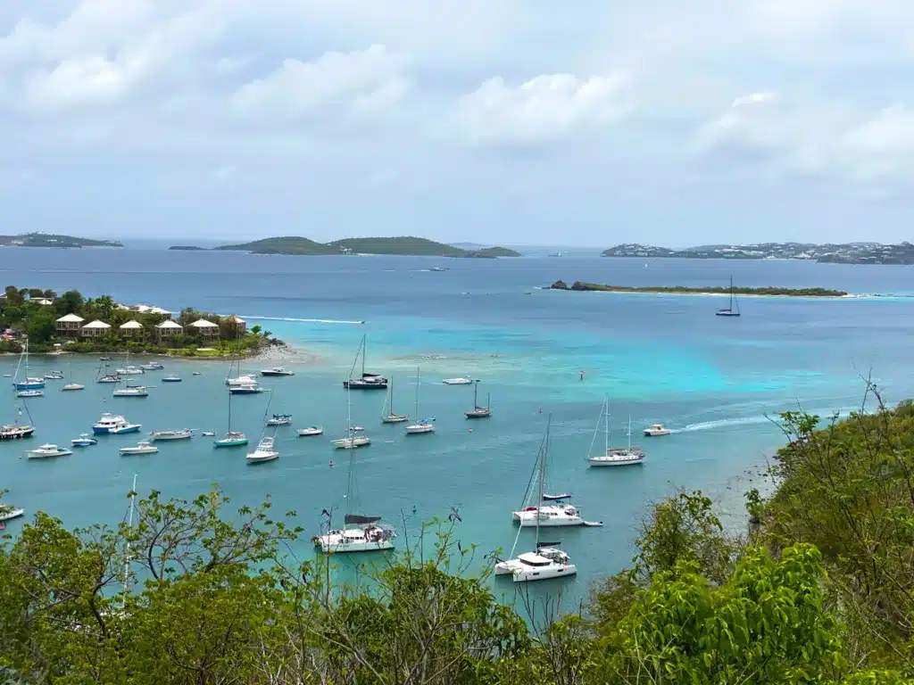 Aerial view of a bay with numerous boats anchored in clear turquoise water, surrounded by lush greenery and distant islands under a cloudy sky.
