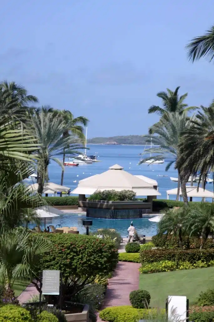 Tropical resort with palm trees, a pool, and cabanas overlooking a bay with boats and yachts anchored, with an island visible in the background under clear blue skies.