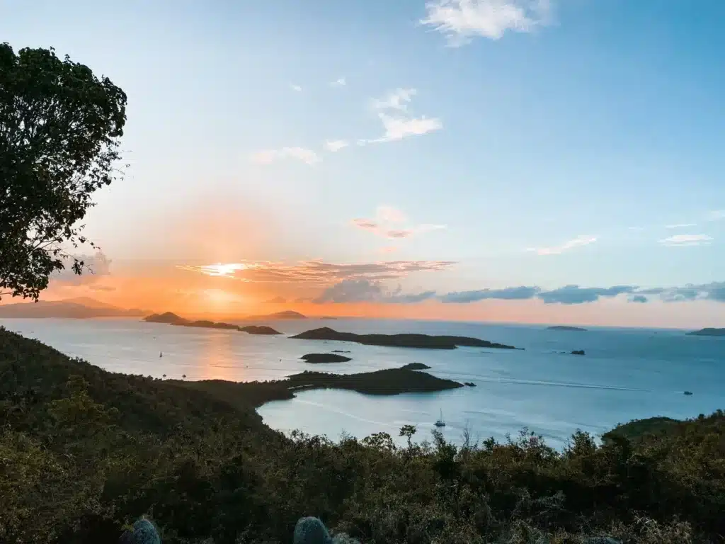 A serene sunset over a tranquil bay with small islands scattered across the water, viewed from a hillside with trees in the foreground and a partly cloudy sky.