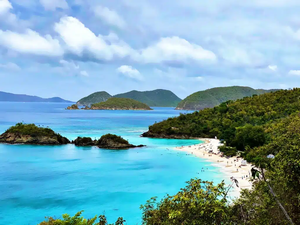 Aerial view of a tropical beach with turquoise waters, lush green hills, scattered small islands, and people enjoying the sandy shoreline under a partly cloudy sky.