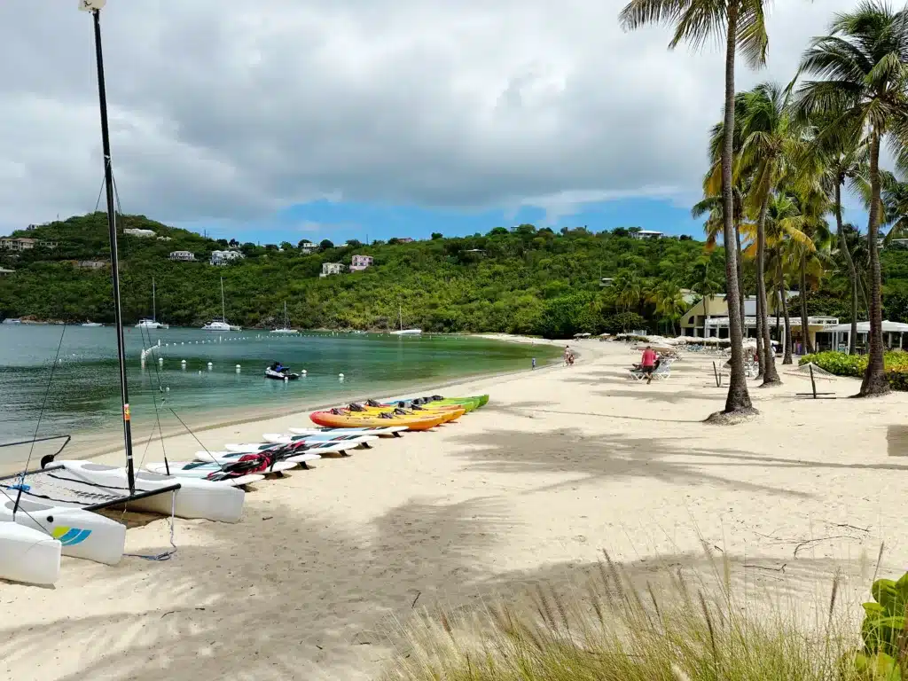 A sandy beach with catamarans, kayaks, and paddleboards lined up. Palm trees are scattered nearby, and hills with houses are in the background under a partly cloudy sky.