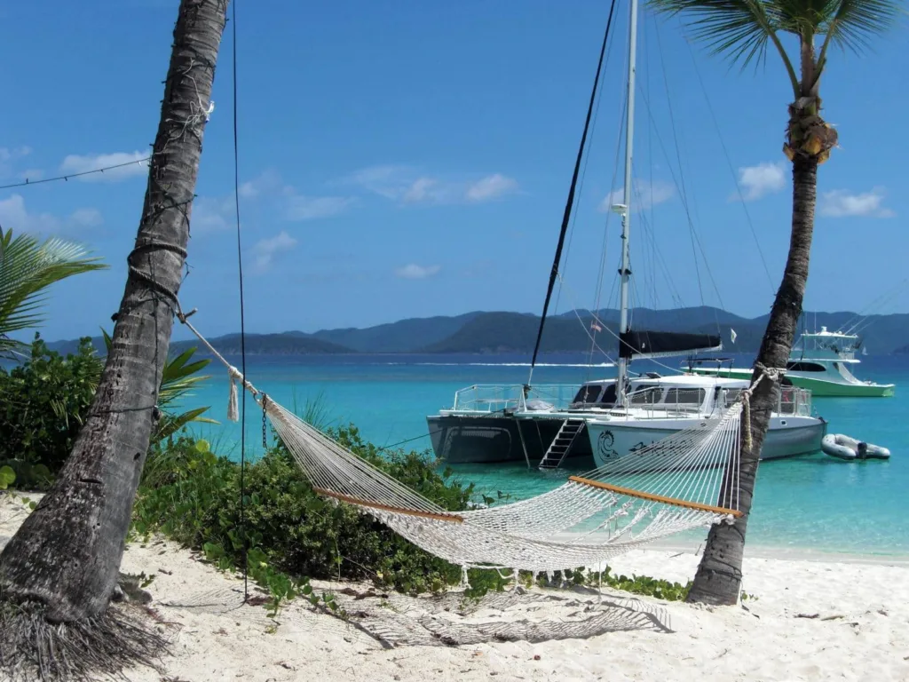 A hammock is tied between two palm trees on a sandy beach with clear blue waters and two sailboats anchored in the background.