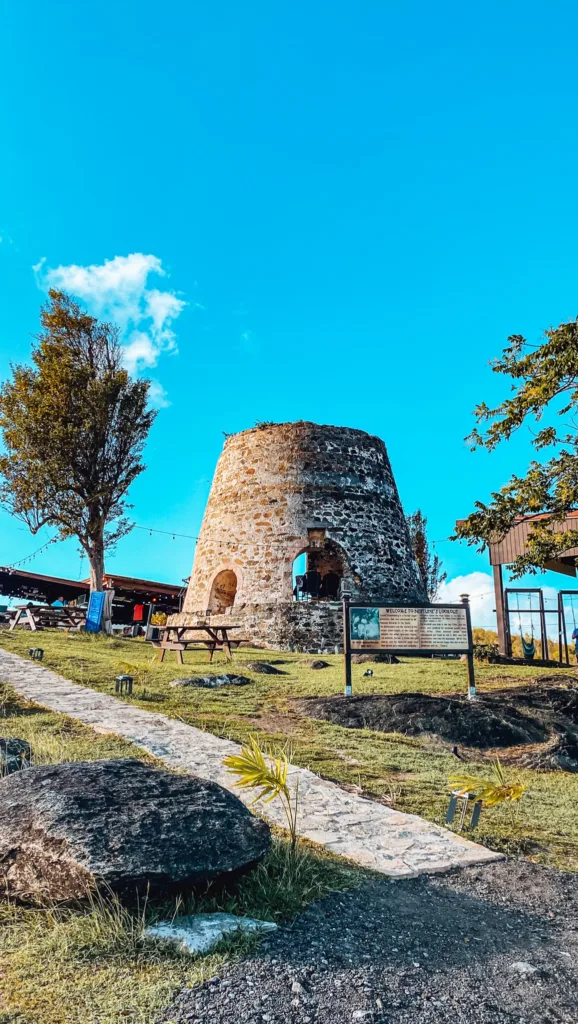 A stone tower with an entrance stands on a grassy area under a bright blue sky, surrounded by picnic tables and a few trees. Information boards and signage are visible nearby.