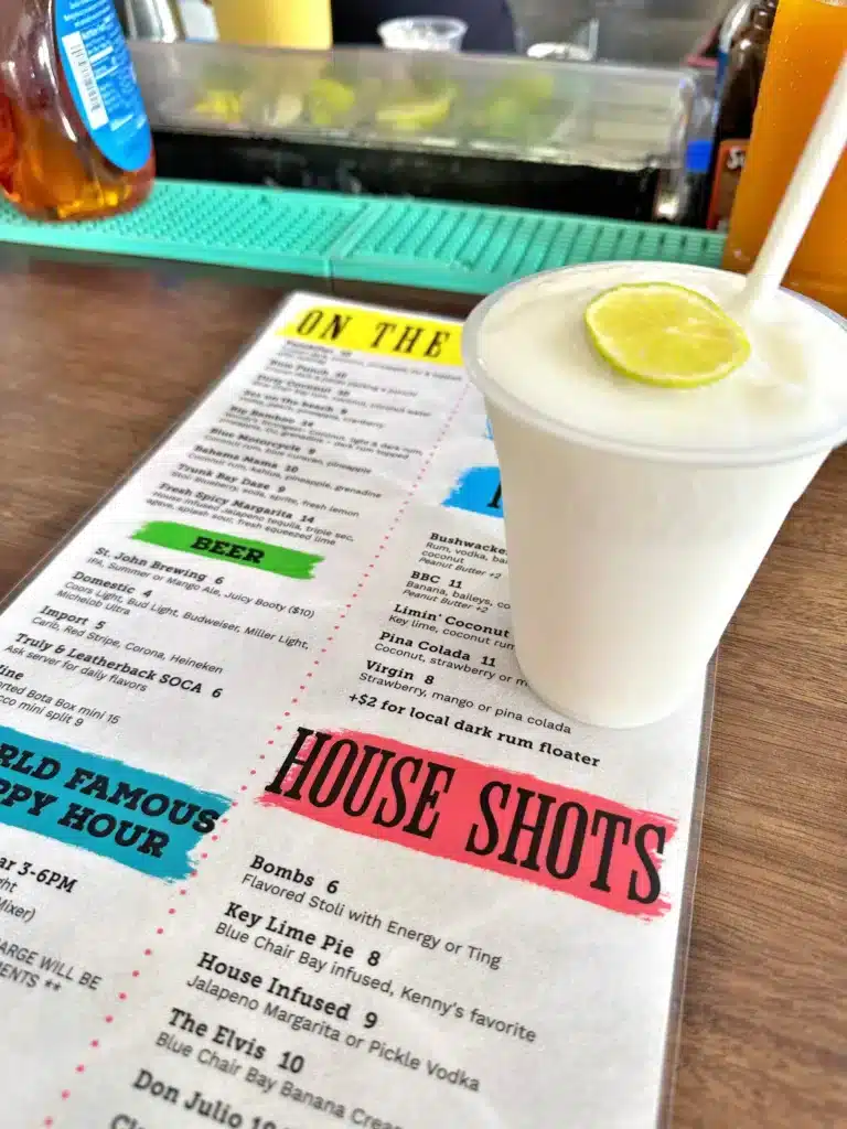 A frozen cocktail garnished with a lime slice sits on a table next to a drink menu titled "House Shots.
