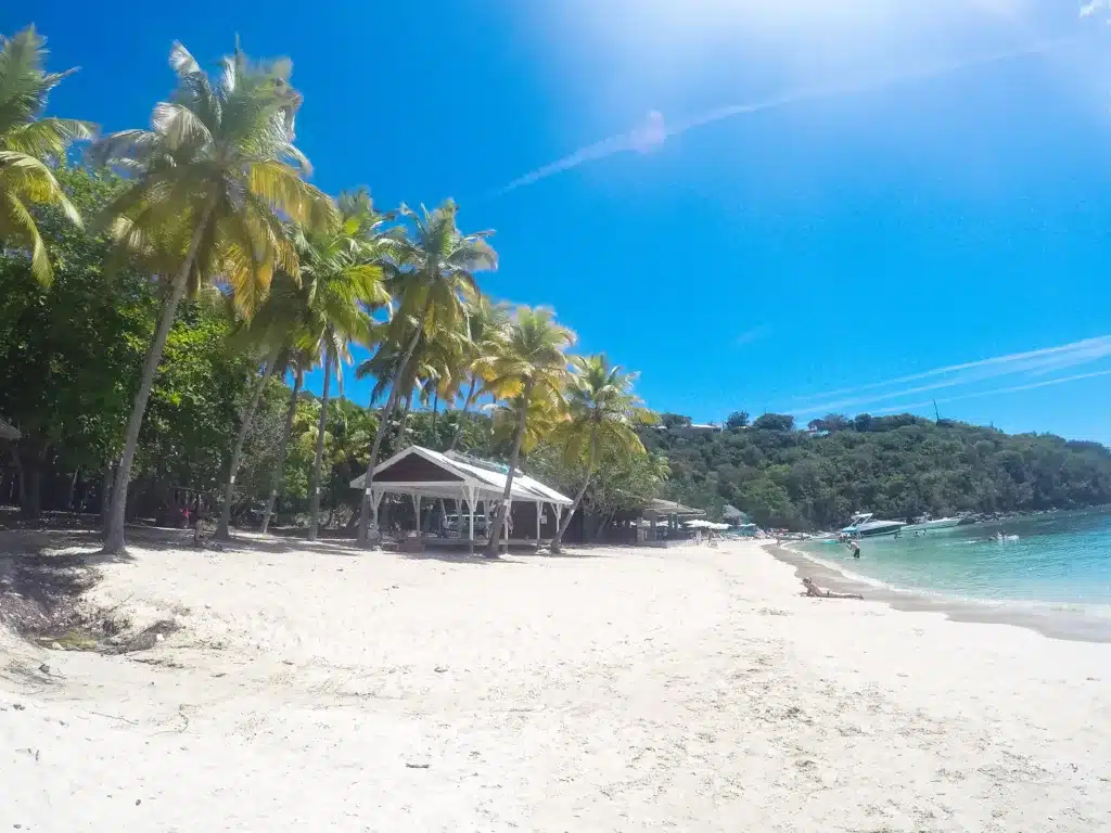 A tropical beach with white sand, clear blue water, and palm trees. A few small boats are docked at the shoreline, and a wooden shelter stands near the trees.