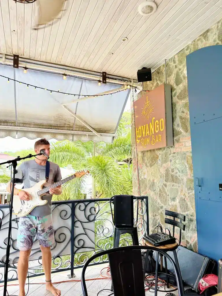 A man plays an electric guitar and sings into a microphone on a small outdoor stage at Lovango Rum Bar, with lights strung overhead and tropical foliage visible in the background.