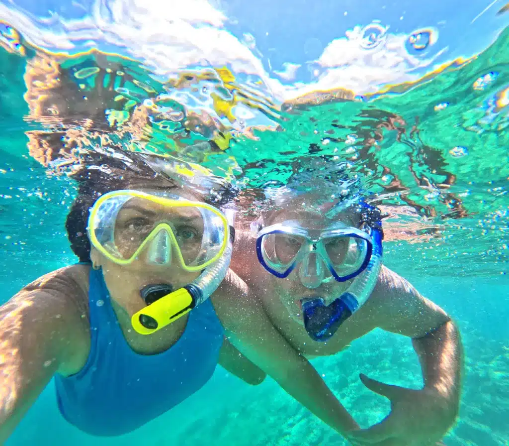Two people wearing snorkel masks swim underwater in clear blue water, with reflections of the sky and clouds visible on the surface above them.