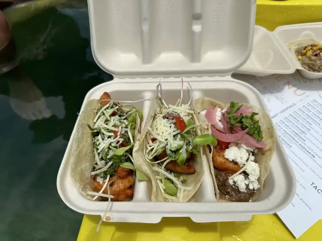Three tacos in an open biodegradable container, with varying toppings including shredded cheese, greens, and pickled onions, placed on a yellow surface with a menu partially visible underneath.