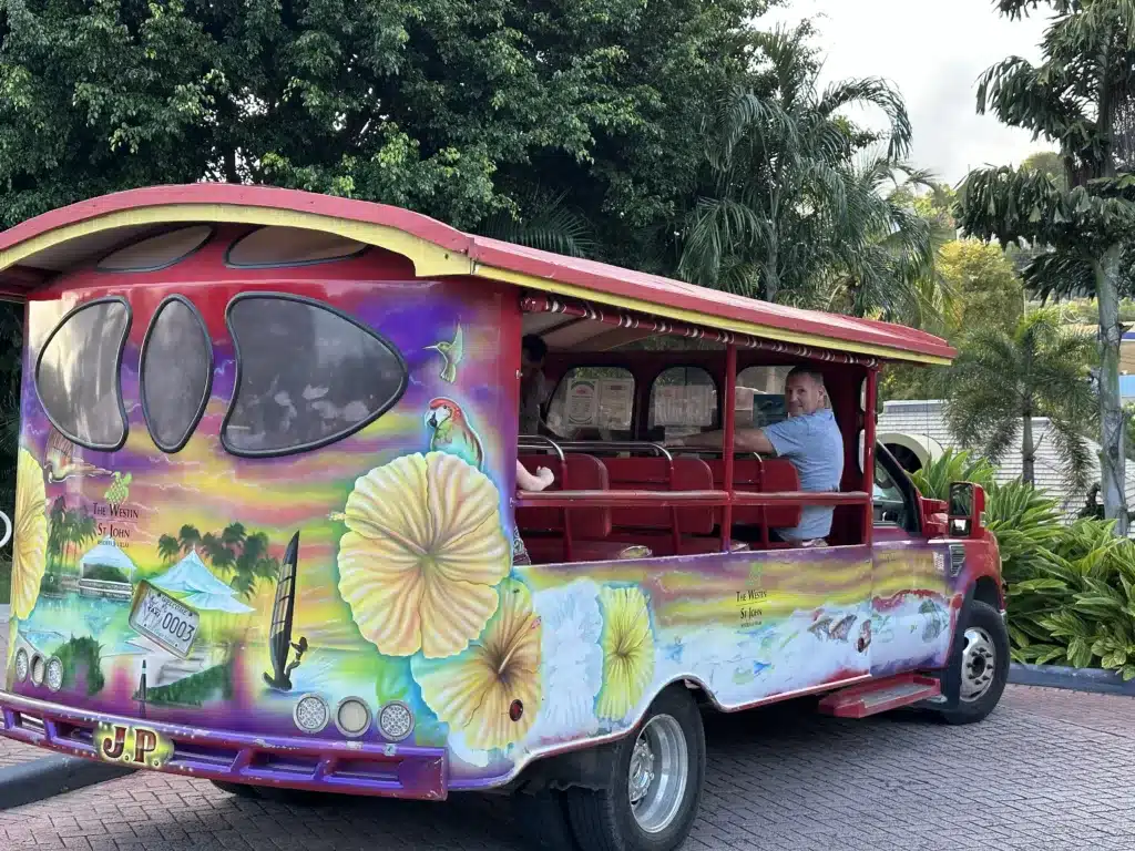 A man in a blue shirt drives a colorful open-sided tour bus with detailed murals. The bus is parked on a brick road with trees and foliage in the background.