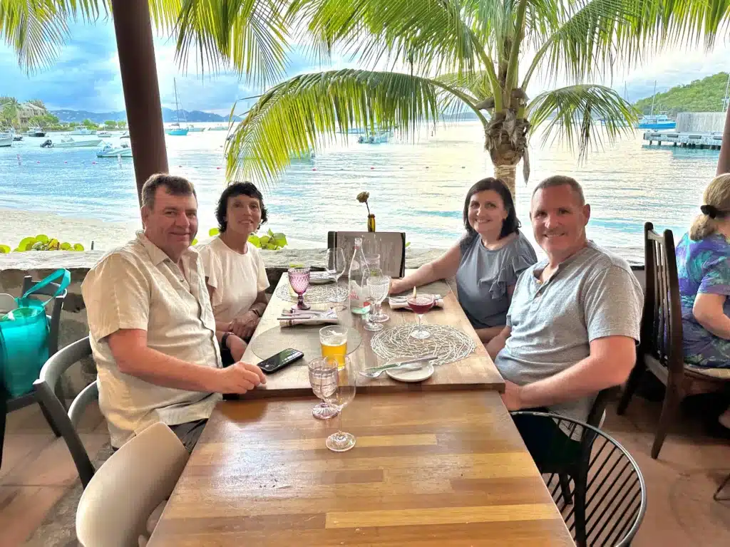 Four people sit around a table with drinks and plates at a seaside restaurant. Palm trees, boats, and a calm sea are visible in the background.