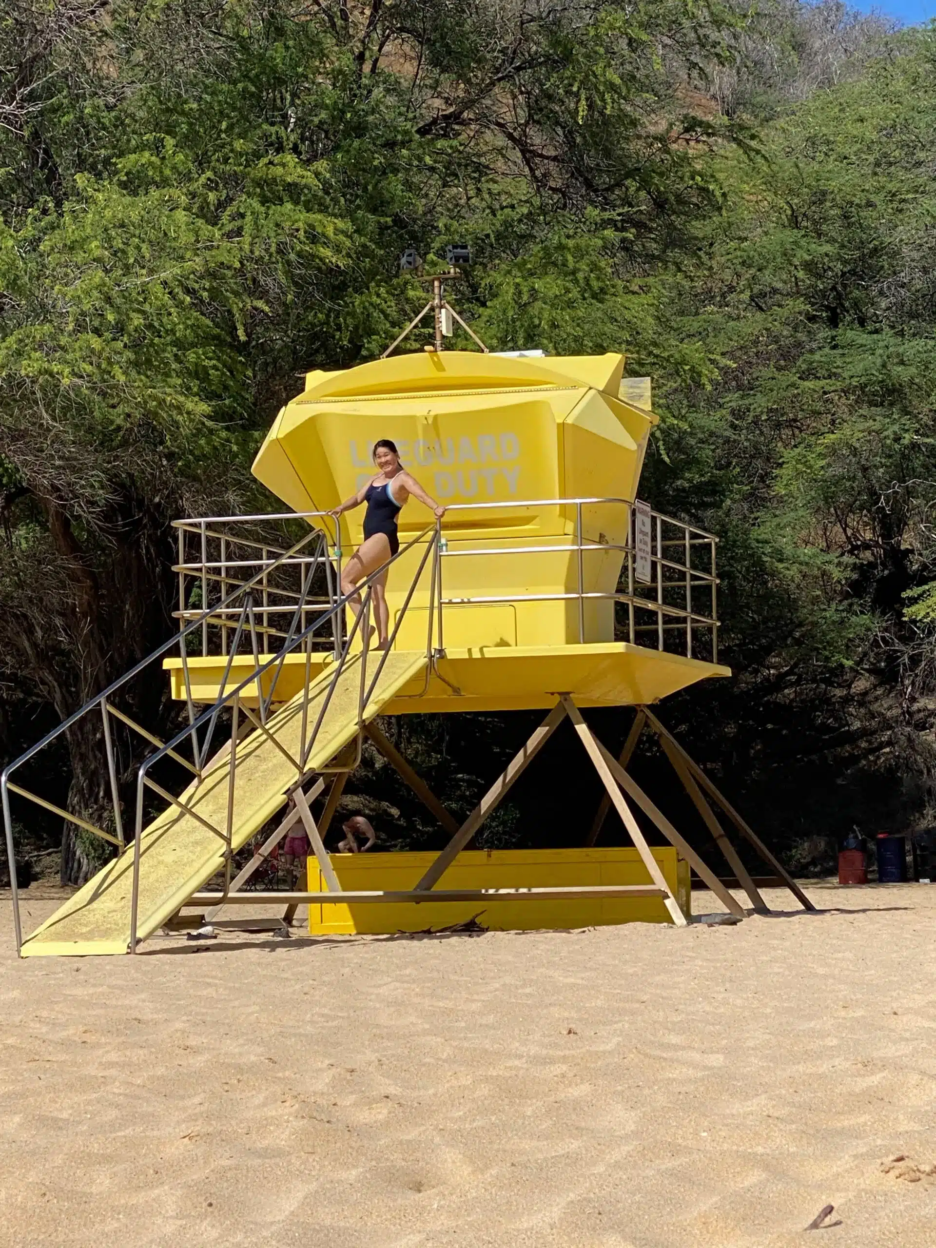 A person in a swimsuit stands on the stairs of a yellow lifeguard tower on a sandy beach with trees in the background.