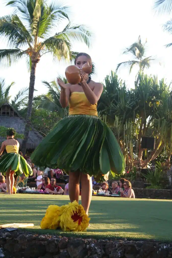 A young woman performs a traditional hula dance, holding a gourd, wearing a green grass skirt and yellow top, with palm trees and an audience in the background.