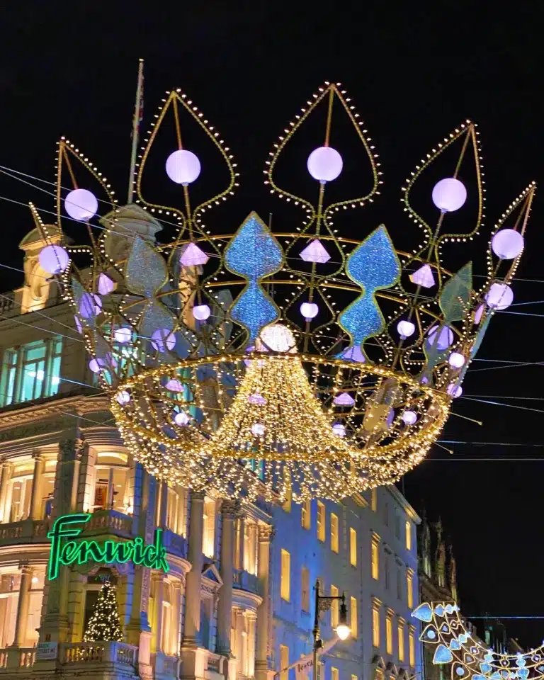 A large illuminated crown-shaped decoration hangs over a street at night, with the Fenwick storefront in the background.