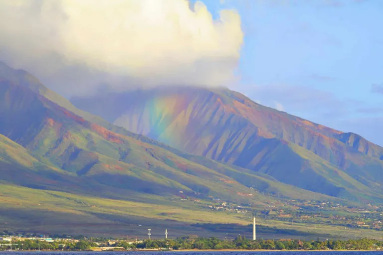 A faint rainbow appears in front of a cloud-covered mountain with green slopes and scattered buildings in the foreground.