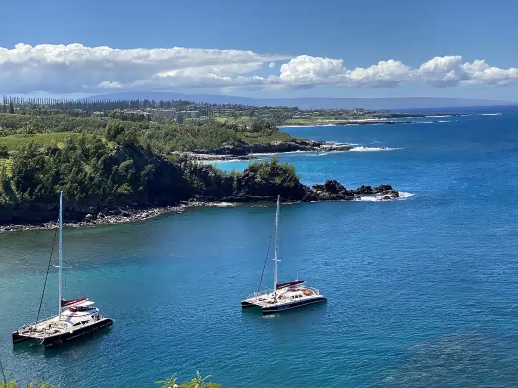 Two catamarans are anchored in a small cove with lush green cliffs and clear blue water, under a partly cloudy sky. The coastline and hills are visible in the background.