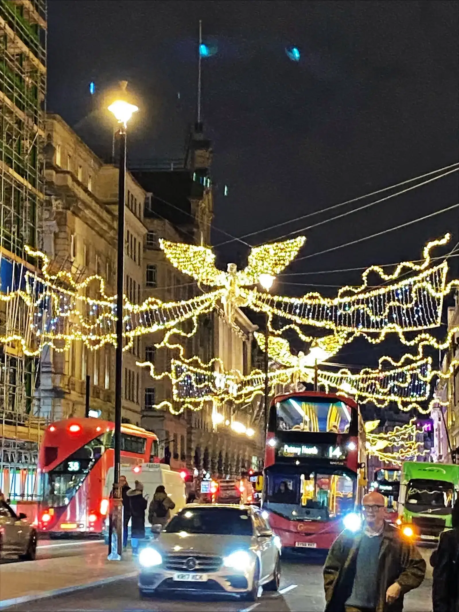 London street at night decorated with glowing angel-shaped holiday lights above red double-decker buses, a highlight for those visiting London during Christmas.