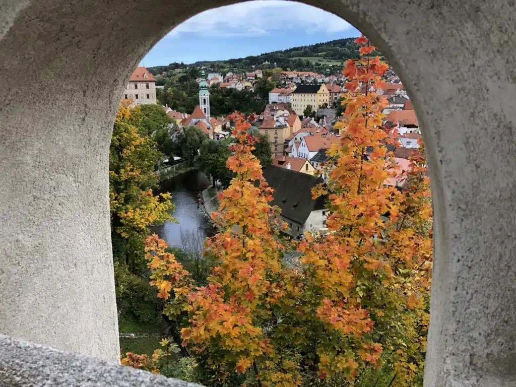 View through an archway showcasing a town with red-roofed buildings, a river, and trees with autumn foliage. The scene includes a mix of greenery and hilly background under a partly cloudy sky.