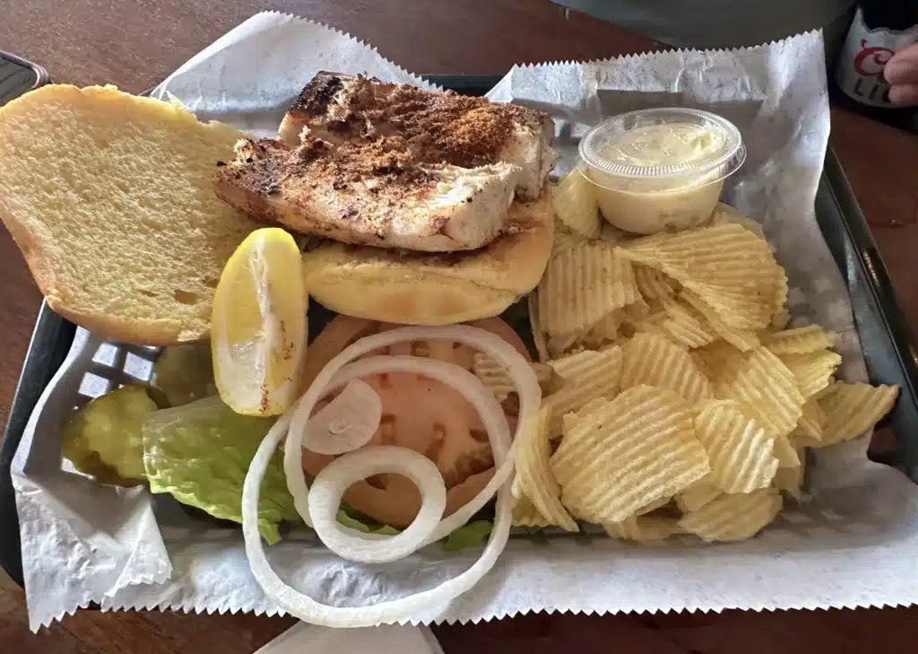 A tray with a fish sandwich on a bun, a lemon wedge, lettuce, sliced tomato, onion rings, pickle slices, a serving of potato chips, and a small container of sauce.