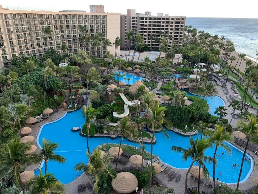 Aerial view of a tropical resort in Kaanapali, showcasing multiple blue swimming pools surrounded by palm trees and lounge chairs, with multi-story hotel buildings in the background and the ocean on the horizon. Discover the best things to do in this paradise.