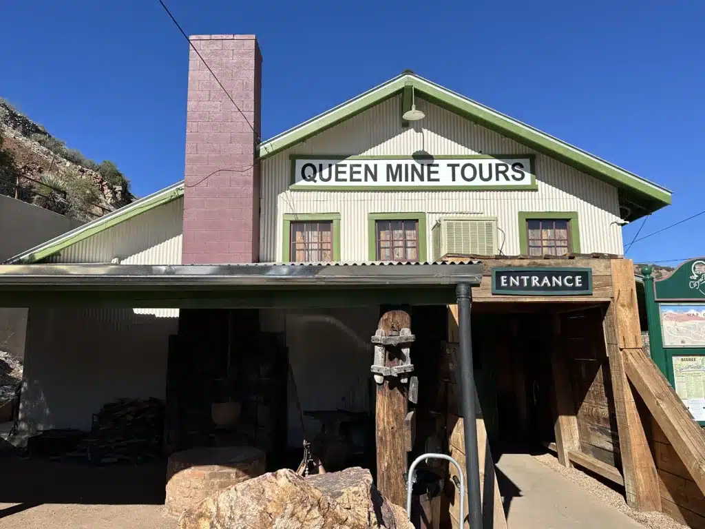 Front view of a building with a sign reading "Queen Mine Tours." A tall chimney stands on the left, with an entrance to the right featuring wooden posts. Ideal for day trips from Tucson, this site invites exploration and history just beyond the city.