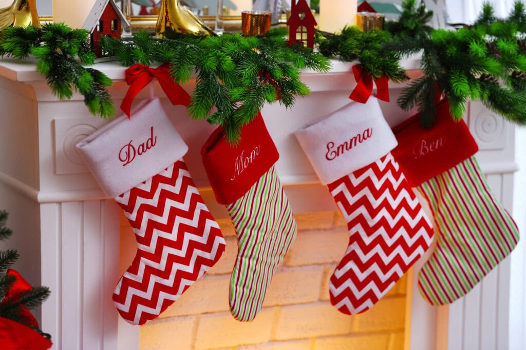 Four personalized Christmas stockings labeled Dad, Mom, Emma, and Ben hang from a garland-decorated white mantel above the fireplace—perfect for holding the best stocking stuffers for travelers in your family.