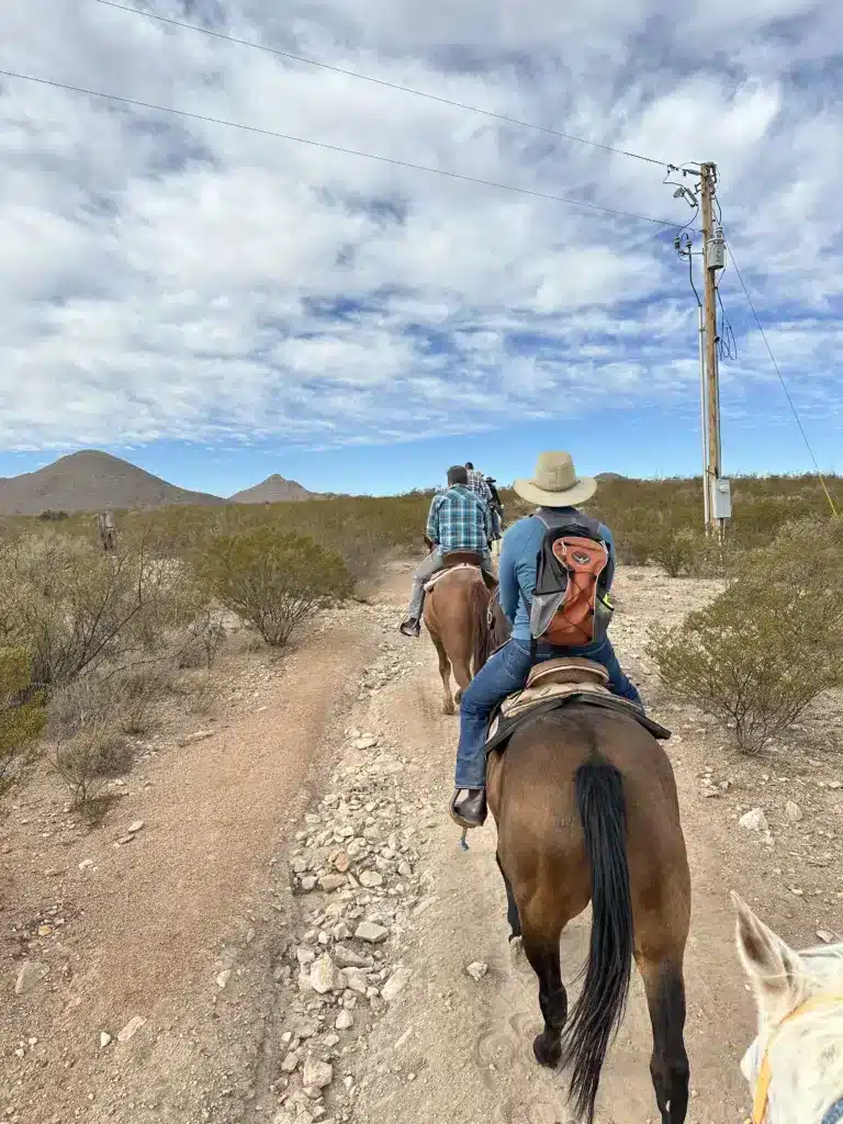 Two people ride horses on a rocky path through a shrub-filled landscape, with clouds in the sky and distant hills, offering a perfect vignette of day trips from Tucson.