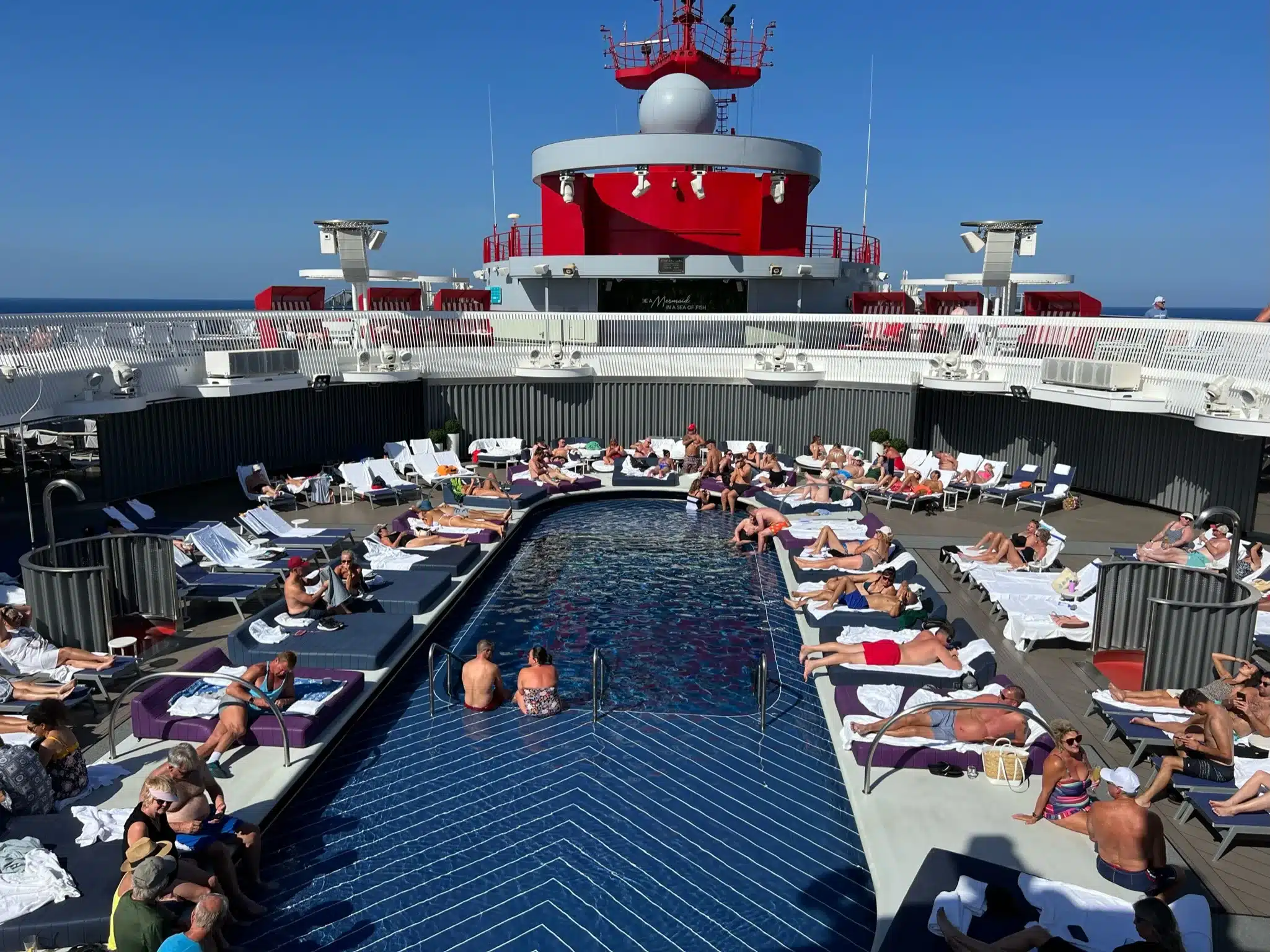 People lounging around a sparkling rectangular pool on a cruise ship deck, under a clear blue sky, enjoy the sunny tranquility. Don’t forget to include swimwear and sunscreen in your cruise packing list for moments like this.