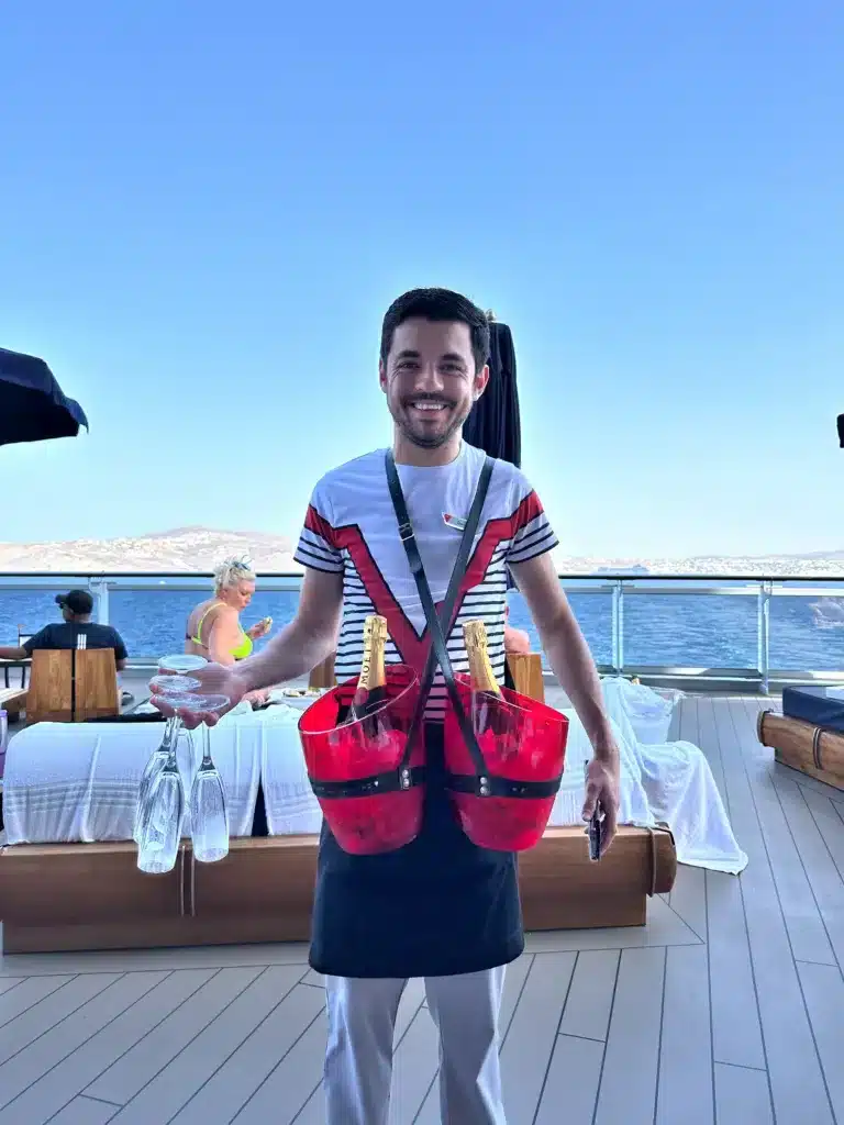 A person stands on a deck, showcasing their well-prepared cruise packing list with glasses and two ice buckets filled with bottles. The ocean glistens in the background, accompanied by lounge chairs and an inviting umbrella nearby.