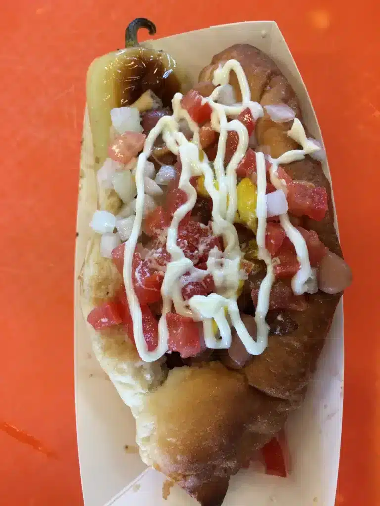A hot dog topped with diced tomatoes, onions, and mayonnaise, nestled in a paper tray on an orange surface—perfect for a quick bite between day trips from Tucson.