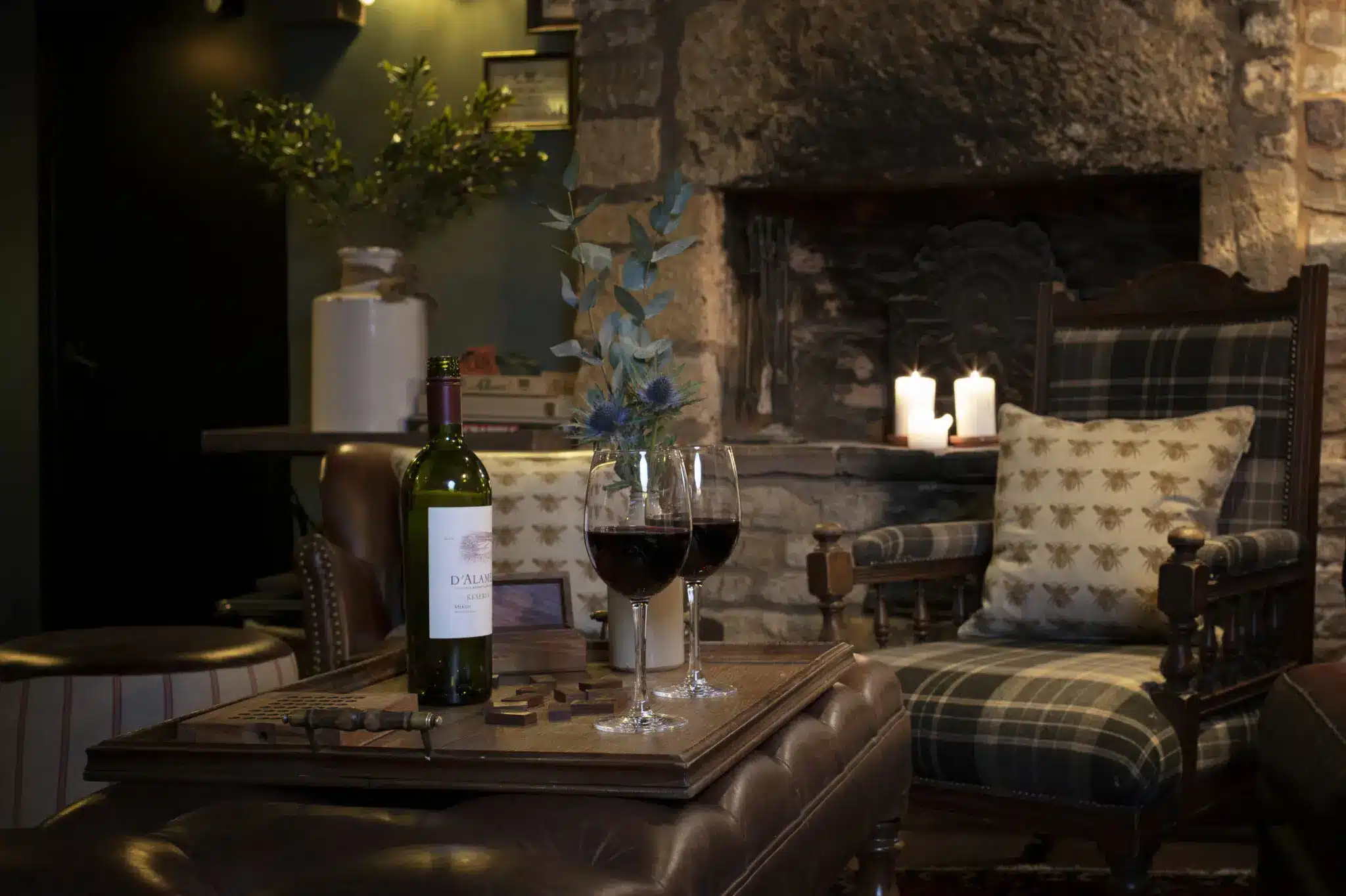 A cozy room reminiscent of the best Cotswold pubs, featuring a stone fireplace, two glasses of red wine, and a bottle on an ottoman. Two armchairs with patterned pillows surround the warmth, complemented by candles and a plant.