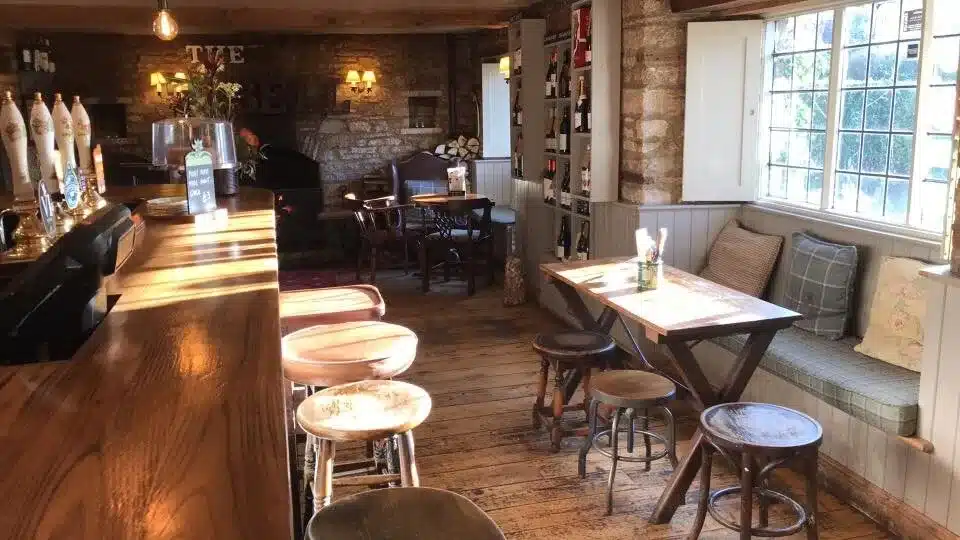 Nestled among the best Cotswold pubs, this cozy interior features wooden floors, a line of barstools at a classic wooden bar, and cushioned seating by a window. Displays of bottles line the walls as soft lighting casts a warm atmosphere.